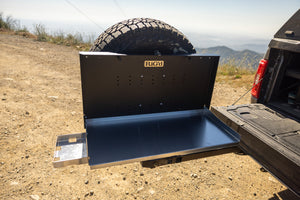 A black outdoor camp table set on a truck with a spare tire in the background.