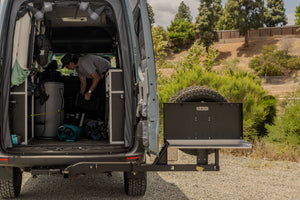 A black camp table mounted on a slide-out platform from a camper van, with a man organizing gear inside the van and a natural outdoor background.