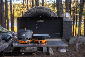 Outdoor camping table setup with a black fuel stove system and two cooking pots, one on a metal table in a wooded forest setting.