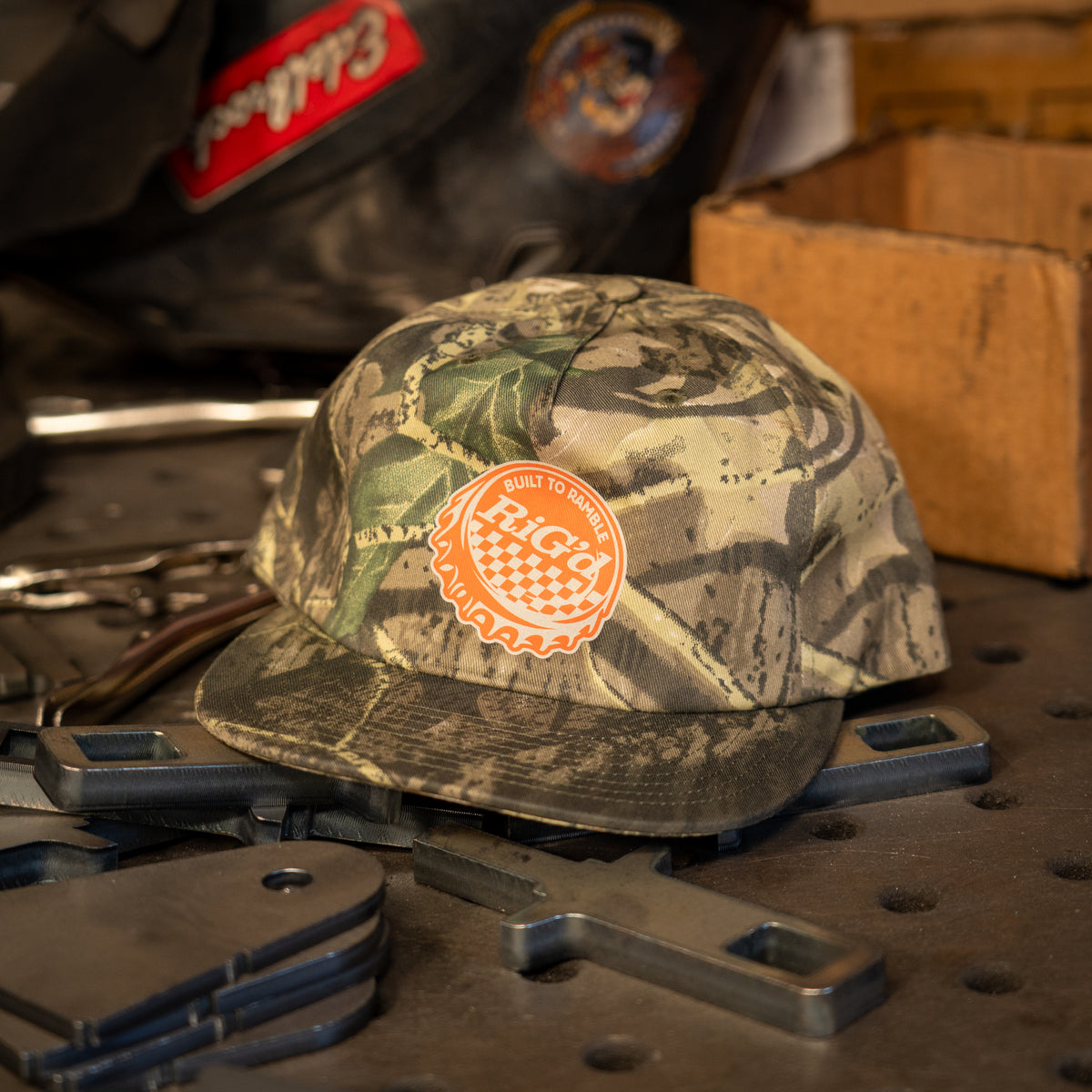 A camo baseball hat with an orange and white bottle cap 'RIGd' logo patch on the front, resting on a rustic wooden surface with a warm, blurred workshop background.