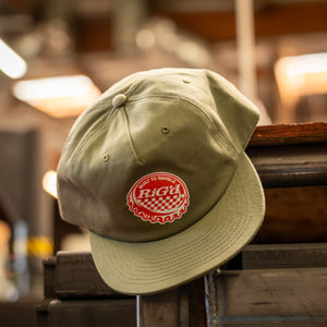 An olive green baseball hat with a red and white bottle cap 'RIGd' logo patch on the front, resting on a rustic wooden surface with a warm, blurred workshop background.