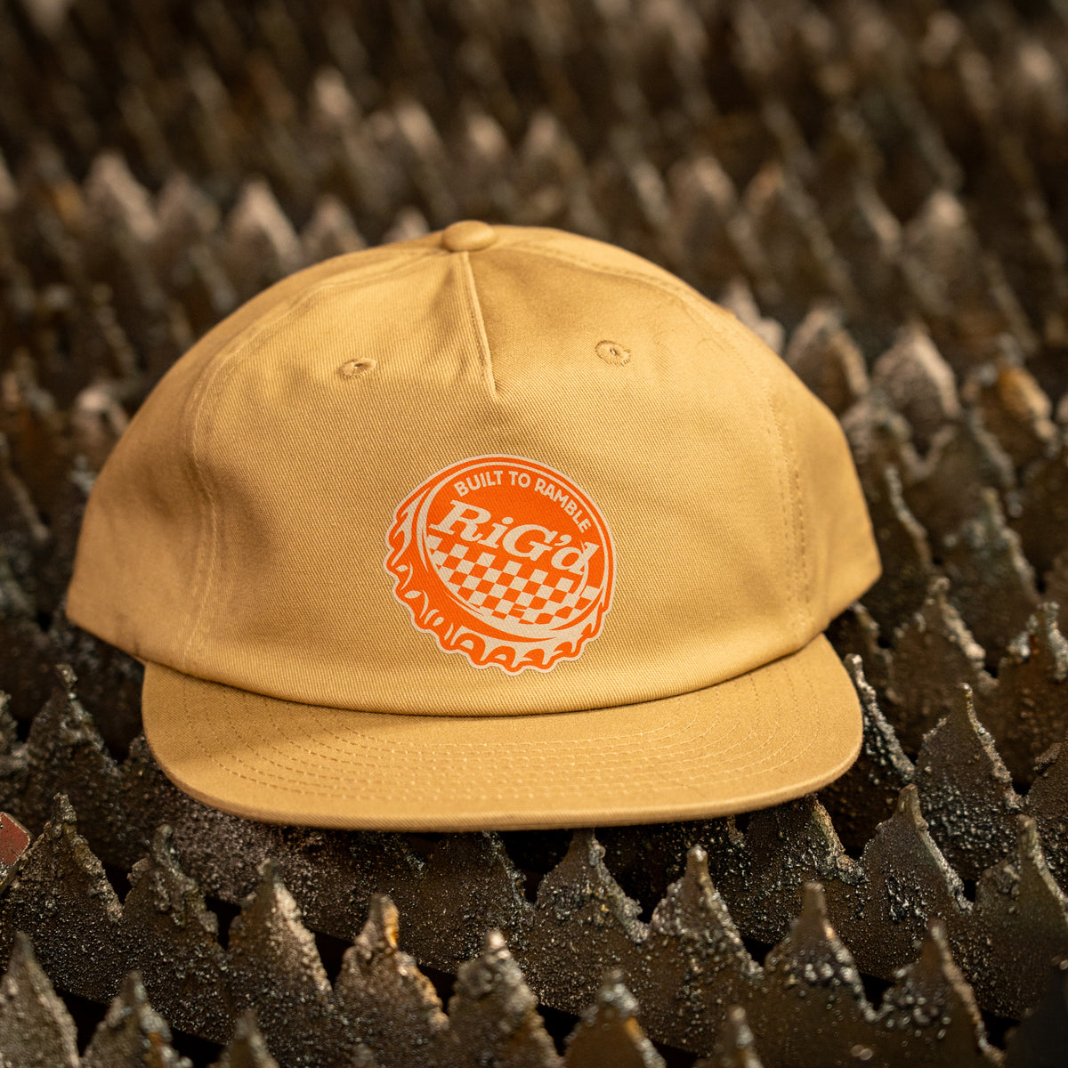 A tan baseball hat with a red and white bottle cap 'RIGd' logo patch on the front, resting on a rustic wooden surface with a warm, blurred workshop background.