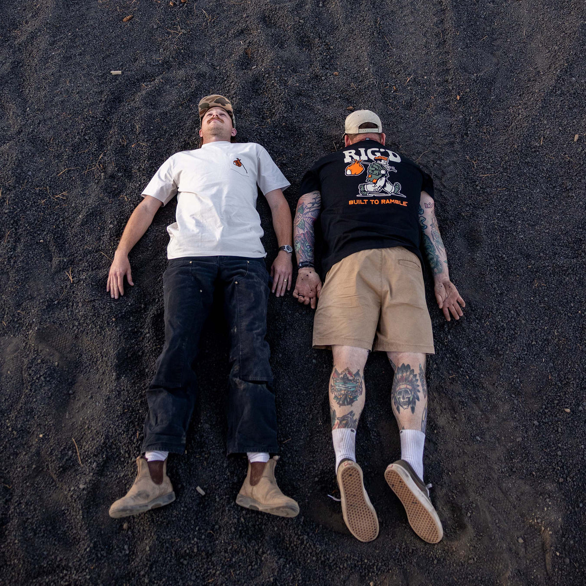 Two people lying on a dark sandy surface, one wearing a white shirt and dark pants, the other in a black shirt with visible tattoos.