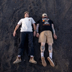 Two people lying on a dark sandy surface, one wearing a white shirt and dark pants, the other in a black shirt with visible tattoos.
