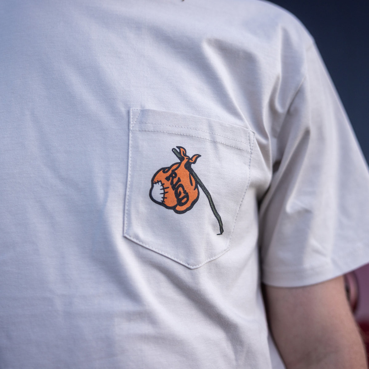 White t-shirt with a small orange and black design on the chest pocket against a dark background