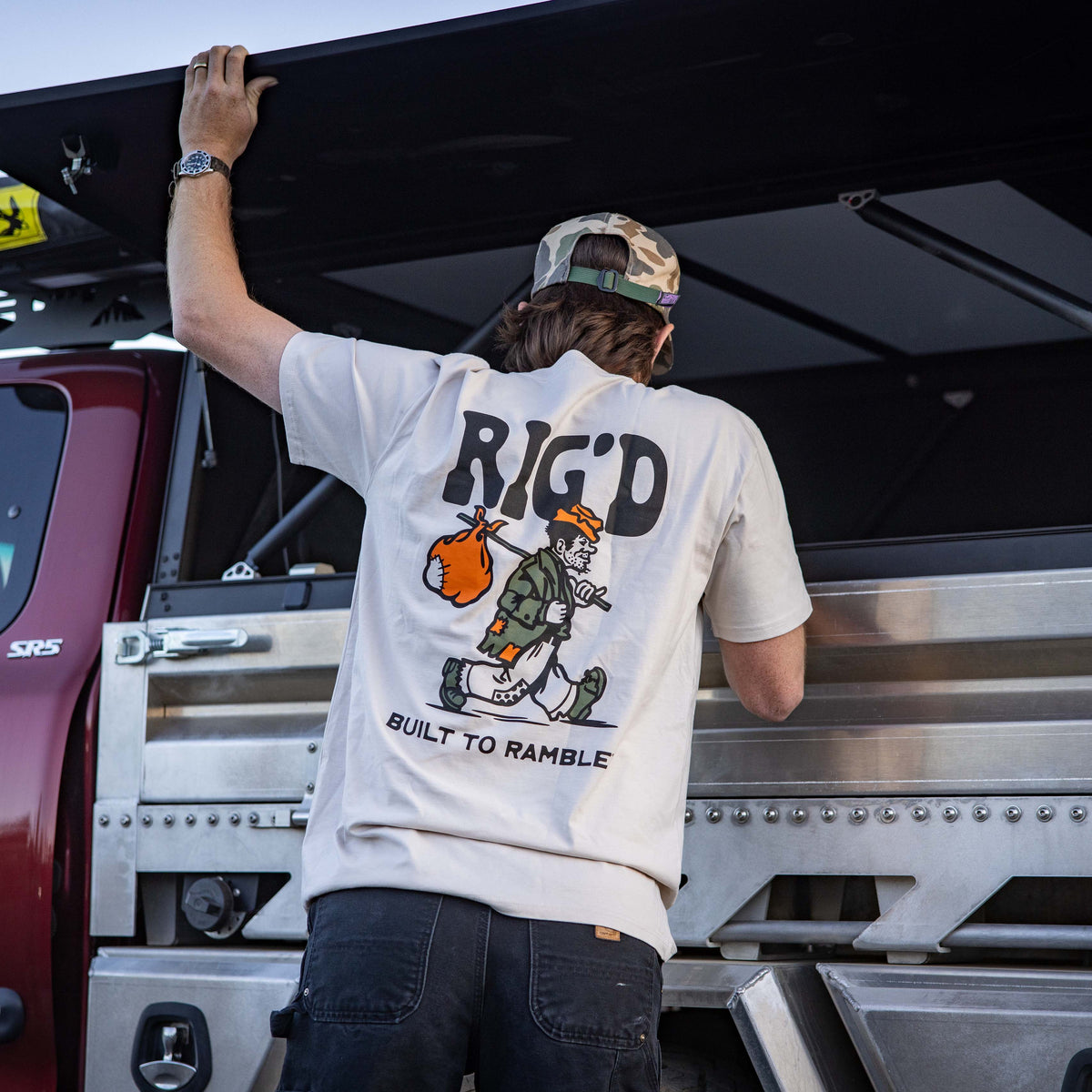 Person wearing a '
RiG'D' t-shirt with a graphic design, standing next to a truck.